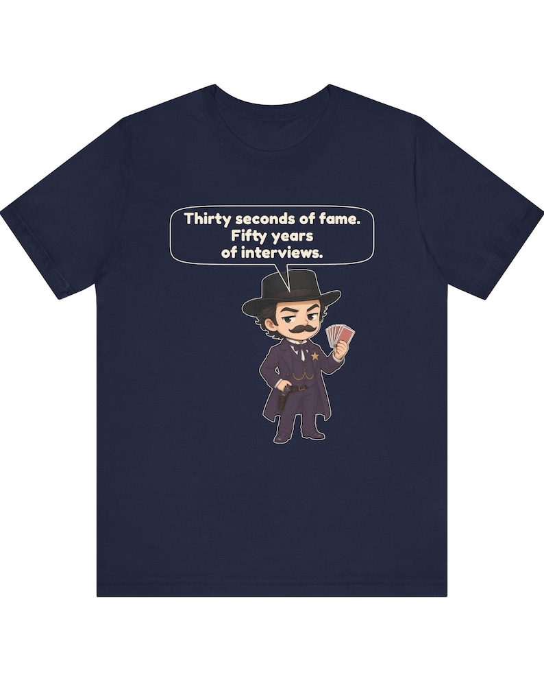 Pu&ograve; includere: Una t-shirt blu navy con un'illustrazione a cartoni animati di un personaggio con baffi, cappello e cappotto neri, che tiene in mano carte da gioco e una pistola. Un fumetto sopra il personaggio contiene il testo: Thirty seconds of fame. Fifty years of interviews.