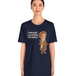 May include: A navy blue short-sleeve t-shirt featuring a cartoon illustration of a character in a brown coat and white scarf holding a quill and candle, with a speech bubble containing the text: Underpaid, overworked, still festive.