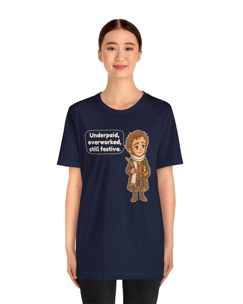May include: A navy blue short-sleeve t-shirt featuring a cartoon illustration of a character in a brown coat and white scarf holding a quill and candle, with a speech bubble containing the text: Underpaid, overworked, still festive.