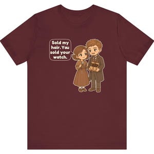 May include: A maroon short-sleeve t-shirt featuring a cartoon illustration of two people in brown clothing. A speech bubble contains the text: Sold my hair. You sold your watch. The characters are holding small gift boxes in their hands.