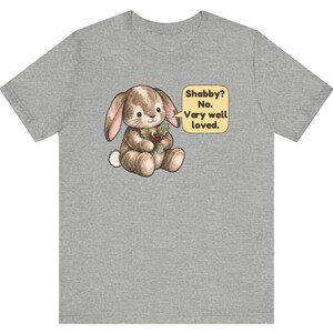 May include: A heather gray t-shirt featuring a brown, illustrated bunny holding a sprig of holly. A yellow speech bubble next to the bunny contains the text: Shabby? No. Very well loved. The bunny has a white tail and stitched details.