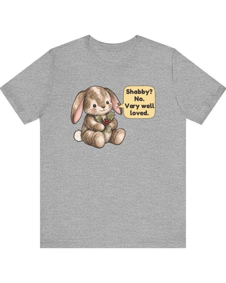 May include: A heather gray t-shirt featuring a brown, illustrated bunny holding a sprig of holly. A yellow speech bubble next to the bunny contains the text: Shabby? No. Very well loved. The bunny has a white tail and stitched details.