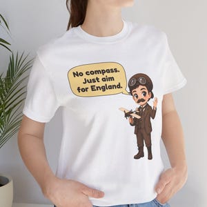 May include: A white t-shirt featuring a cartoon illustration of a pilot in a brown suit, cap, and goggles holding a small airplane. A speech bubble above the pilot contains the text: No compass. Just aim for England.