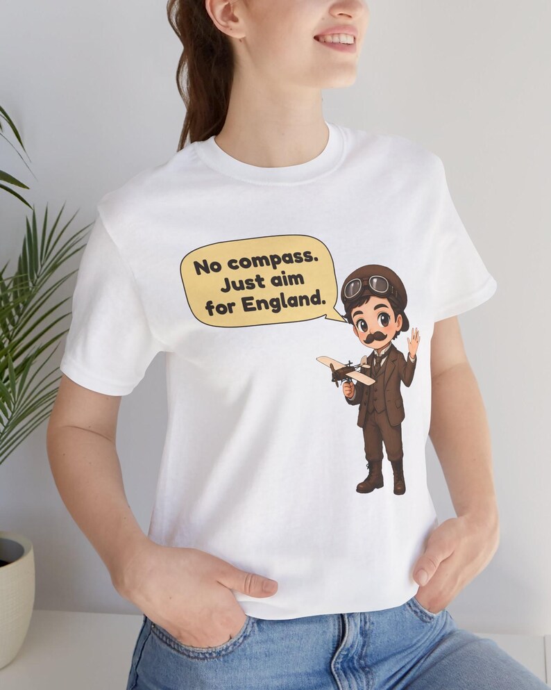 May include: A white t-shirt featuring a cartoon illustration of a pilot in a brown suit, cap, and goggles holding a small airplane. A speech bubble above the pilot contains the text: No compass. Just aim for England.