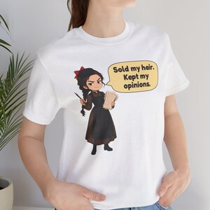 May include: A white t-shirt featuring a cartoon illustration of a person with a dark braid, a red hair bow, and a brown dress with a black apron. The character holds a quill and paper, with a speech bubble that reads: Sold my hair. Kept my opinions.