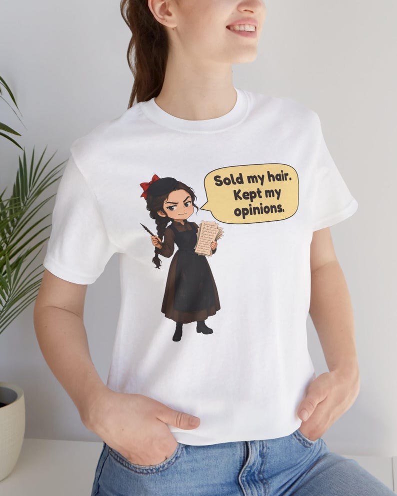 May include: A white t-shirt featuring a cartoon illustration of a person with a dark braid, a red hair bow, and a brown dress with a black apron. The character holds a quill and paper, with a speech bubble that reads: Sold my hair. Kept my opinions.