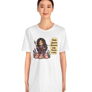 May include: A white t-shirt featuring a graphic of a dark-haired character in a purple dress holding a staff and a small pot, surrounded by three small pigs. A speech bubble contains the text: Rude guests become pigs. House rules.
