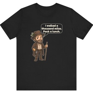 May include: A black t-shirt featuring a cartoon illustration of a bearded hiker wearing a brown hat with a pine sprig, a brown coat, and holding a wooden walking stick. A speech bubble contains the text: I walked a thousand miles. Pack a lunch.