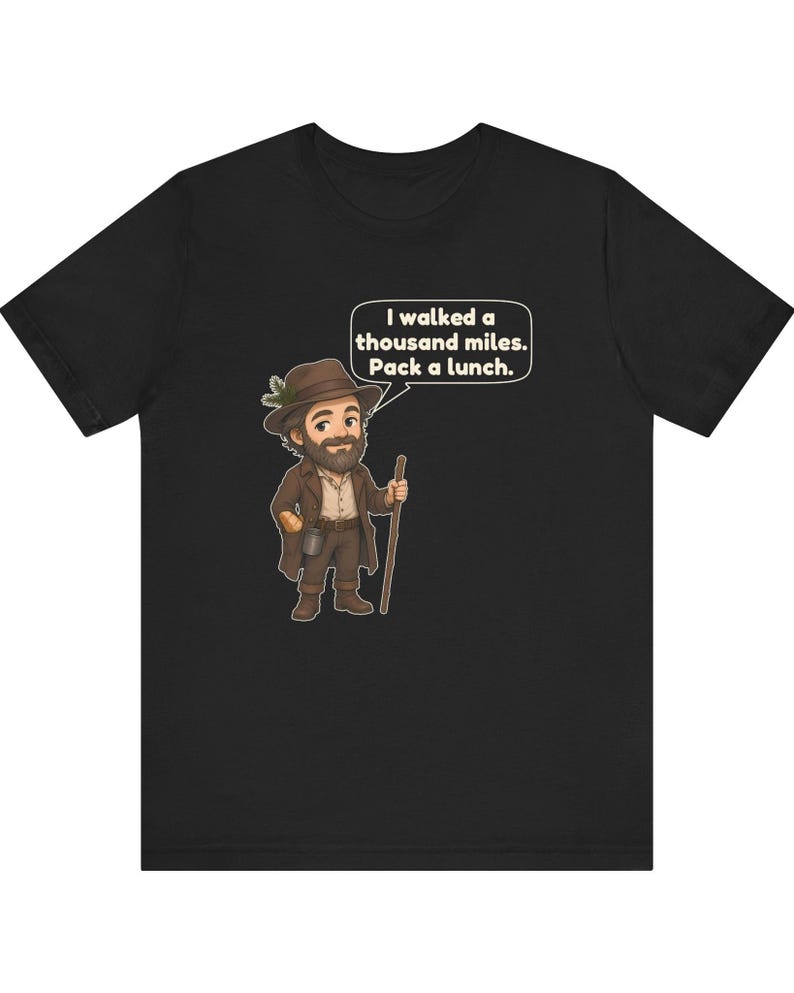 May include: A black t-shirt featuring a cartoon illustration of a bearded hiker wearing a brown hat with a pine sprig, a brown coat, and holding a wooden walking stick. A speech bubble contains the text: I walked a thousand miles. Pack a lunch.
