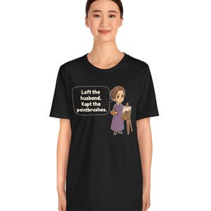 May include: A black t-shirt featuring a cartoon illustration of a person in a purple dress standing next to an easel with a painting. A speech bubble contains the text: Left the husband. Kept the paintbrushes. The design is centered on the chest.