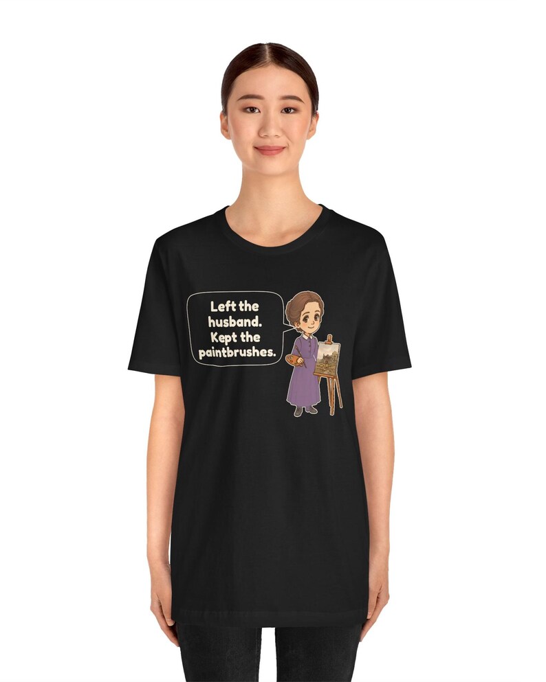 May include: A black t-shirt featuring a cartoon illustration of a person in a purple dress standing next to an easel with a painting. A speech bubble contains the text: Left the husband. Kept the paintbrushes. The design is centered on the chest.