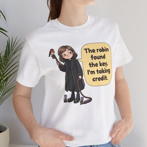 May include: A white t-shirt featuring a cartoon illustration of a person in a black coat holding a large key with a robin perched on it. A yellow speech bubble contains the text: The robin found the key. I'm taking credit.