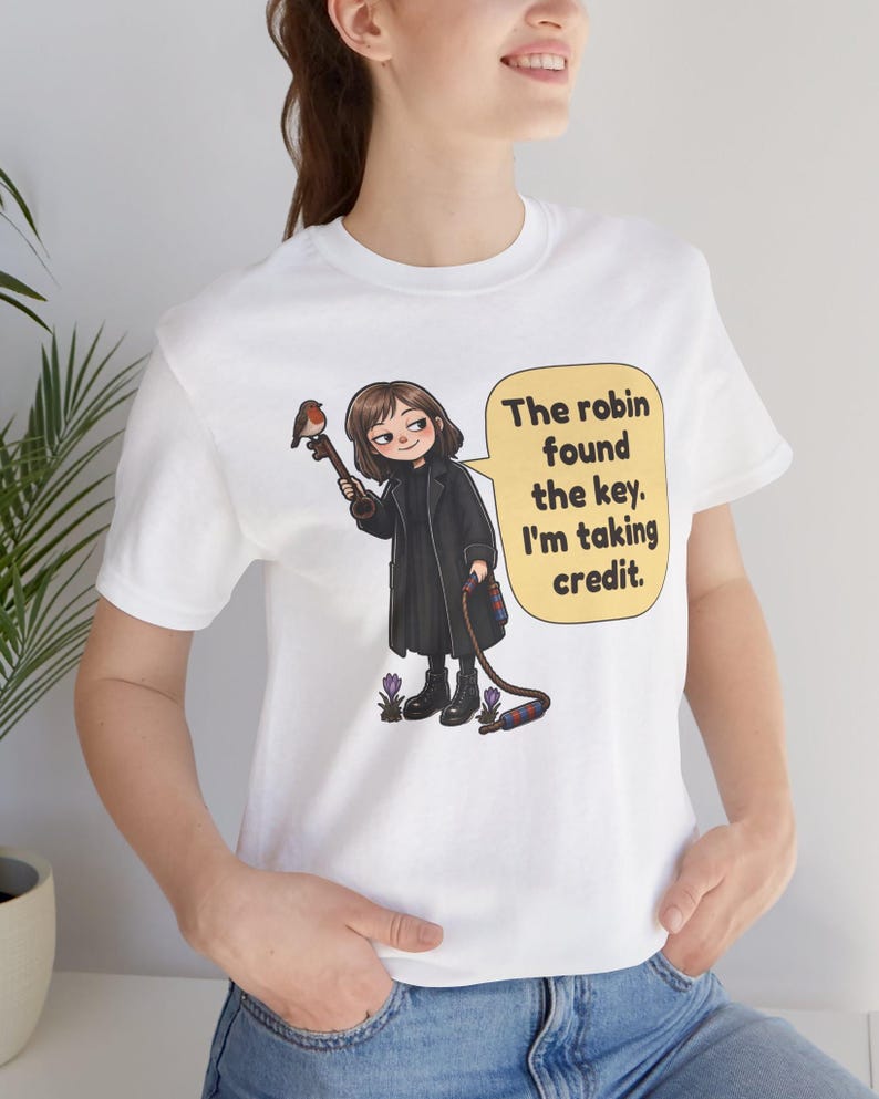 May include: A white t-shirt featuring a cartoon illustration of a person in a black coat holding a large key with a robin perched on it. A yellow speech bubble contains the text: The robin found the key. I'm taking credit.