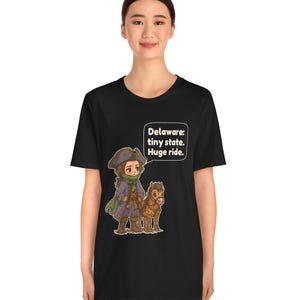 May include: A black t-shirt featuring a cartoon illustration of a person in a historical tricorn hat and coat standing next to a small brown horse. A speech bubble above them contains the text: Delaware: tiny state. Huge ride.