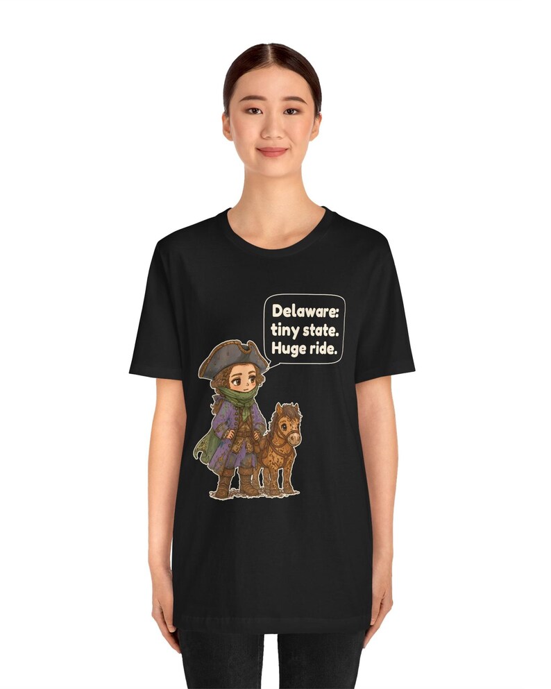 May include: A black t-shirt featuring a cartoon illustration of a person in a historical tricorn hat and coat standing next to a small brown horse. A speech bubble above them contains the text: Delaware: tiny state. Huge ride.