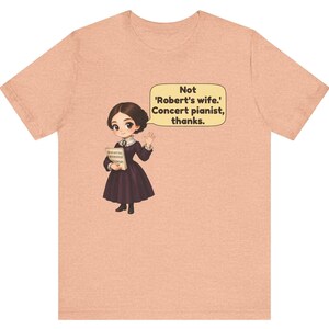 Puede incluir: Una camiseta color melocot&oacute;n con una ilustraci&oacute;n de dibujos animados de una persona con un vestido morado oscuro sosteniendo partituras. Un bocadillo contiene el texto: Not 'Robert's wife.' Concert pianist, thanks.