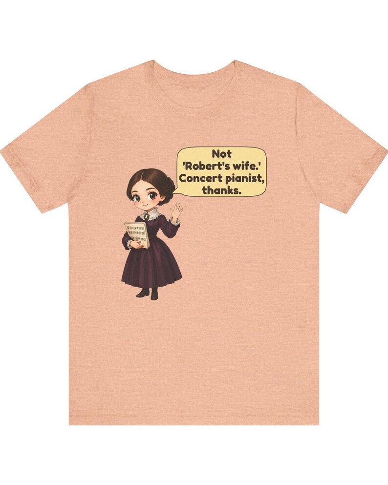 Puede incluir: Una camiseta color melocot&oacute;n con una ilustraci&oacute;n de dibujos animados de una persona con un vestido morado oscuro sosteniendo partituras. Un bocadillo contiene el texto: Not 'Robert's wife.' Concert pianist, thanks.