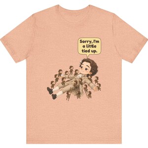 Op de afbeelding: Een perzikkleurig t-shirt met een afbeelding van een groot figuur dat ligt, omringd door vele kleine figuurtjes die touwen en ladders gebruiken om het figuur vast te binden. Een tekstballon bevat de tekst, Sorry, I'm a little tied up.