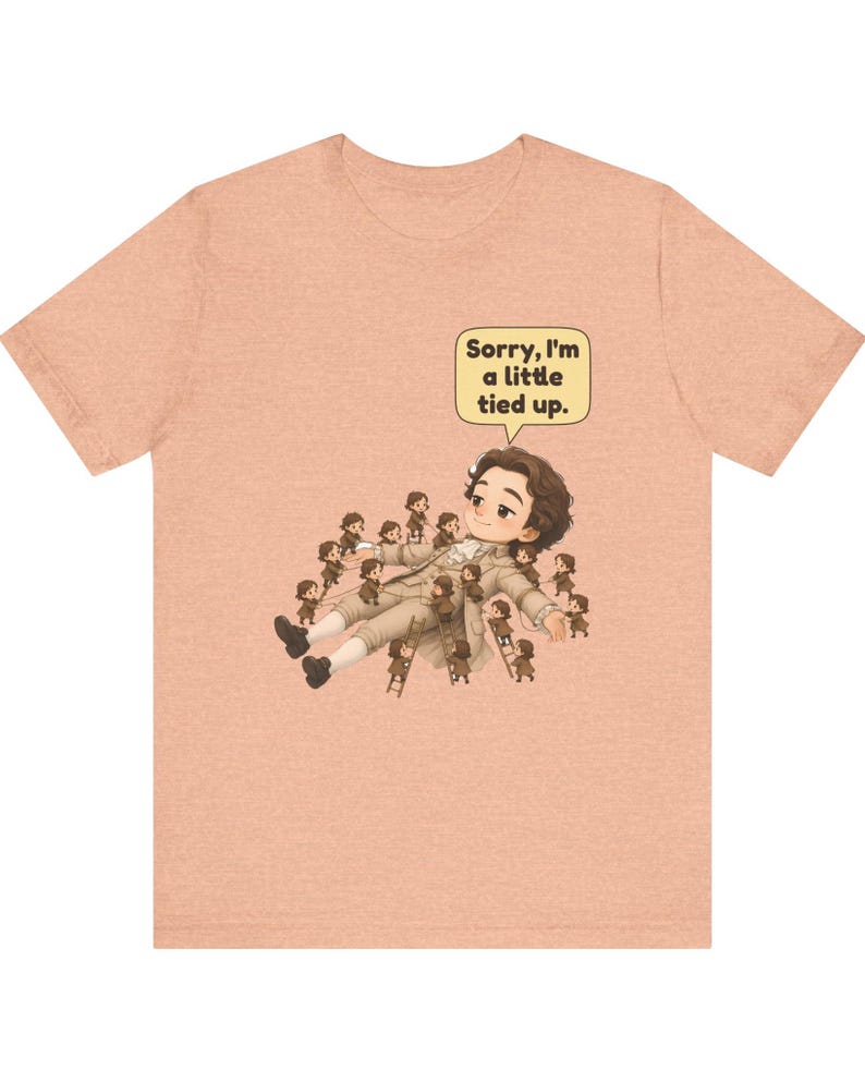 Op de afbeelding: Een perzikkleurig t-shirt met een afbeelding van een groot figuur dat ligt, omringd door vele kleine figuurtjes die touwen en ladders gebruiken om het figuur vast te binden. Een tekstballon bevat de tekst, Sorry, I'm a little tied up.
