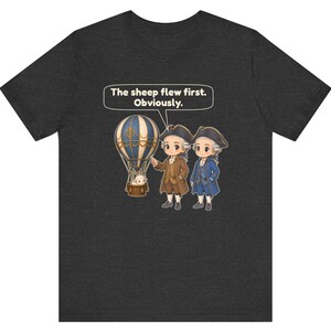 May include: A dark heather gray t-shirt featuring a graphic of two historical figures in tricorn hats and a hot air balloon carrying a sheep. A speech bubble above the figures contains the text, The sheep flew first. Obviously.
