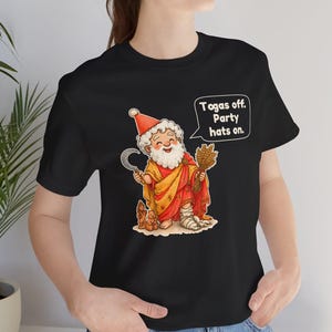 May include: A black t-shirt featuring a cartoon illustration of a bearded figure wearing a red party hat, a yellow and red robe, and a bandage on one foot. The figure holds a sickle and wheat, with a speech bubble that reads: "Togas off. Party hats on."