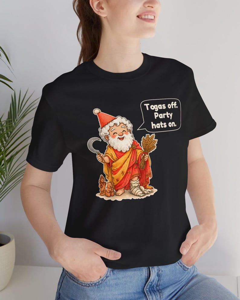 May include: A black t-shirt featuring a cartoon illustration of a bearded figure wearing a red party hat, a yellow and red robe, and a bandage on one foot. The figure holds a sickle and wheat, with a speech bubble that reads: "Togas off. Party hats on."