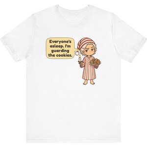 Op de afbeelding: Een wit t-shirt met korte mouwen met een cartoonillustratie van een persoon in een rood-wit gestreepte pyjama en een bijpassende slaapmuts. De persoon houdt een brandende kaars en een bord met koekjes vast, met een tekstballon met de tekst, Everyone's asleep. I'm guarding the cookies.