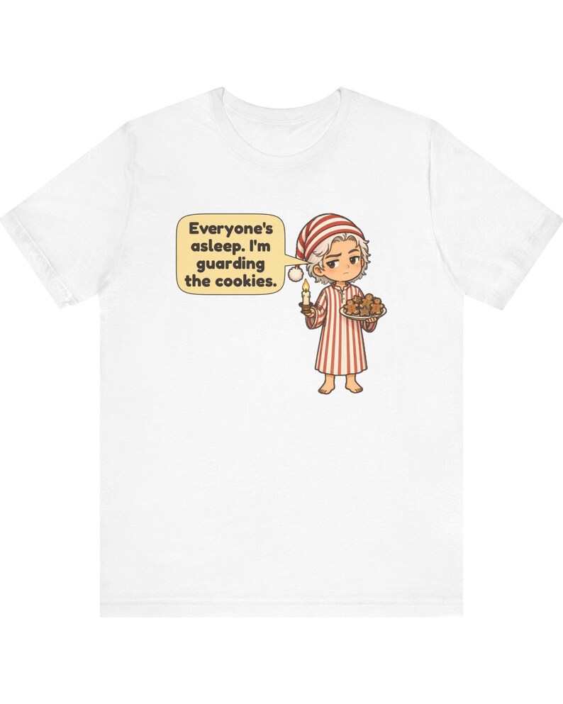 Op de afbeelding: Een wit t-shirt met korte mouwen met een cartoonillustratie van een persoon in een rood-wit gestreepte pyjama en een bijpassende slaapmuts. De persoon houdt een brandende kaars en een bord met koekjes vast, met een tekstballon met de tekst, Everyone's asleep. I'm guarding the cookies.