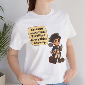 May include: A white t-shirt featuring a cartoon illustration of a historical figure in a blue coat and tricorn hat holding a shovel and compass. A yellow speech bubble contains the text: Arrived uninvited. Fortified everything anyway.