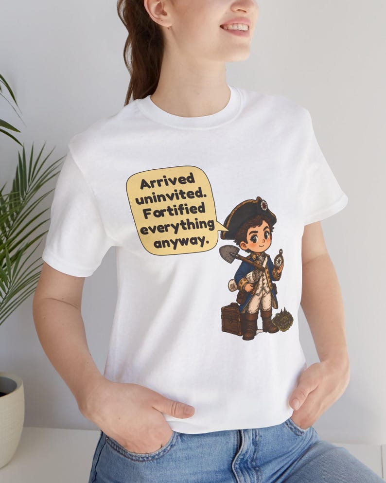 May include: A white t-shirt featuring a cartoon illustration of a historical figure in a blue coat and tricorn hat holding a shovel and compass. A yellow speech bubble contains the text: Arrived uninvited. Fortified everything anyway.