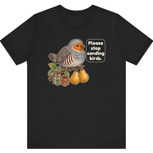 K&ouml;nnte beinhalten: Ein schwarzes T-Shirt mit einer Grafik eines grauen und orangefarbenen Vogels, der auf einem Ast mit zwei Birnen und zwei kleinen Geschenkkartons sitzt. Eine Sprechblase neben dem Vogel enth&auml;lt den Text: "Please stop sending birds."