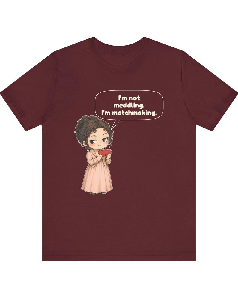 K&ouml;nnte beinhalten: Ein kastanienbraunes T-Shirt mit einer Cartoon-Illustration einer Person mit dunklem, lockigem Haar, die ein hellrosa Kleid und eine Jacke tr&auml;gt und zwei rote Herzen h&auml;lt. Eine Sprechblase dar&uuml;ber enth&auml;lt den Text: I'm not meddling. I'm matchmaking.