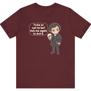 以下が含まれることがあります： 暗い色の服を着た人物が頭蓋骨を持っている漫画風のイラストが描かれた、マルーン色の半袖Tシャツ。吹き出しには「To be or not to be? Ask me again in Act 5.」というテキストが含まれています。デザインは胸の中央に配置されています。