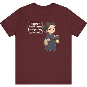 May include: A maroon t-shirt featuring a cartoon illustration of a person in a dark blue dress carrying a satchel and a lit lantern. A speech bubble next to the character contains the text: Retire? At 59 I was just getting started.