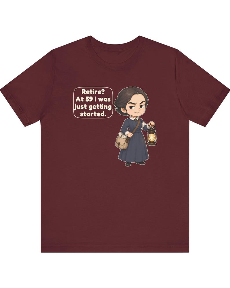 May include: A maroon t-shirt featuring a cartoon illustration of a person in a dark blue dress carrying a satchel and a lit lantern. A speech bubble next to the character contains the text: Retire? At 59 I was just getting started.