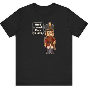 Pu&ograve; includere: Una t-shirt nera con un cartone animato raffigurante uno schiaccianoci in uniforme rossa con dettagli dorati. Un fumetto accanto al personaggio contiene il testo: Hard to crack. Easy to love. Il disegno &egrave; centrato sul davanti.