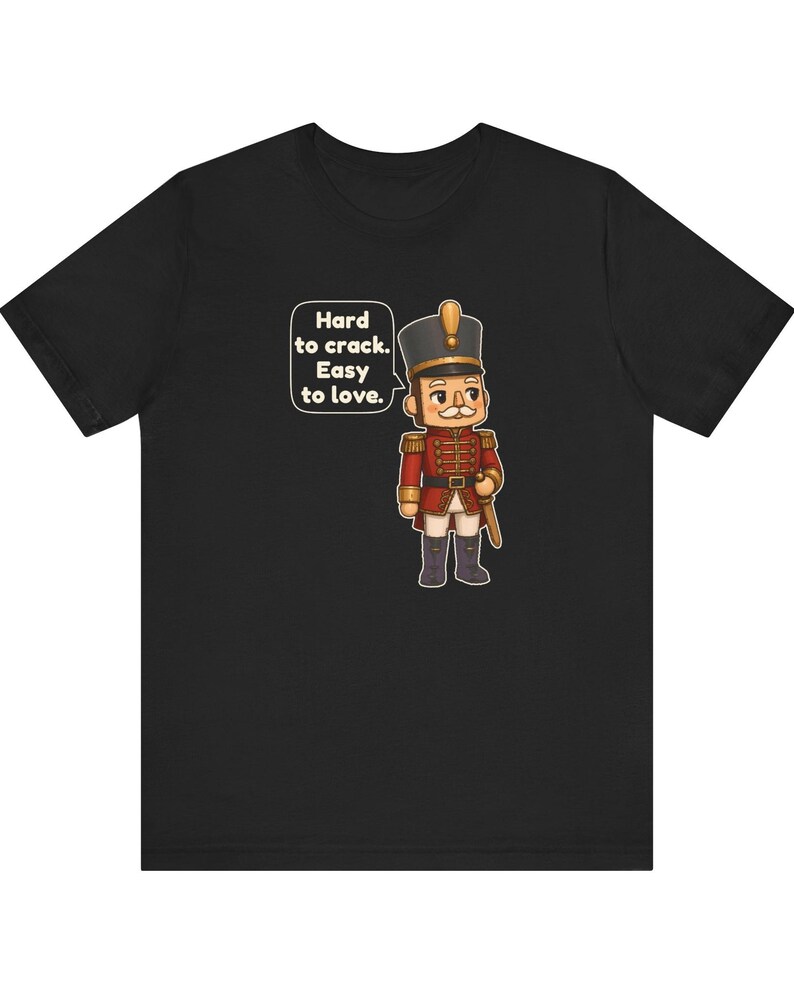 Pu&ograve; includere: Una t-shirt nera con un cartone animato raffigurante uno schiaccianoci in uniforme rossa con dettagli dorati. Un fumetto accanto al personaggio contiene il testo: Hard to crack. Easy to love. Il disegno &egrave; centrato sul davanti.