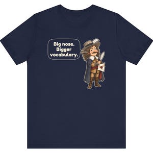 May include: A navy blue short-sleeve t-shirt featuring a cartoon illustration of a historical figure with a large nose, wearing a hat and cape, holding a quill and letter. A speech bubble contains the text: Big nose. Bigger vocabulary.