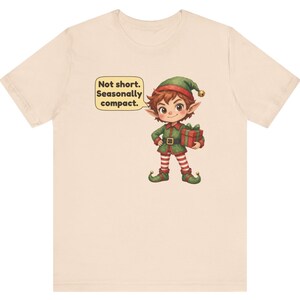 May include: A light beige t-shirt featuring a cartoon elf wearing a green outfit with red and white striped leggings, holding a red gift box. A speech bubble next to the elf contains the text: Not short. Seasonally compact.