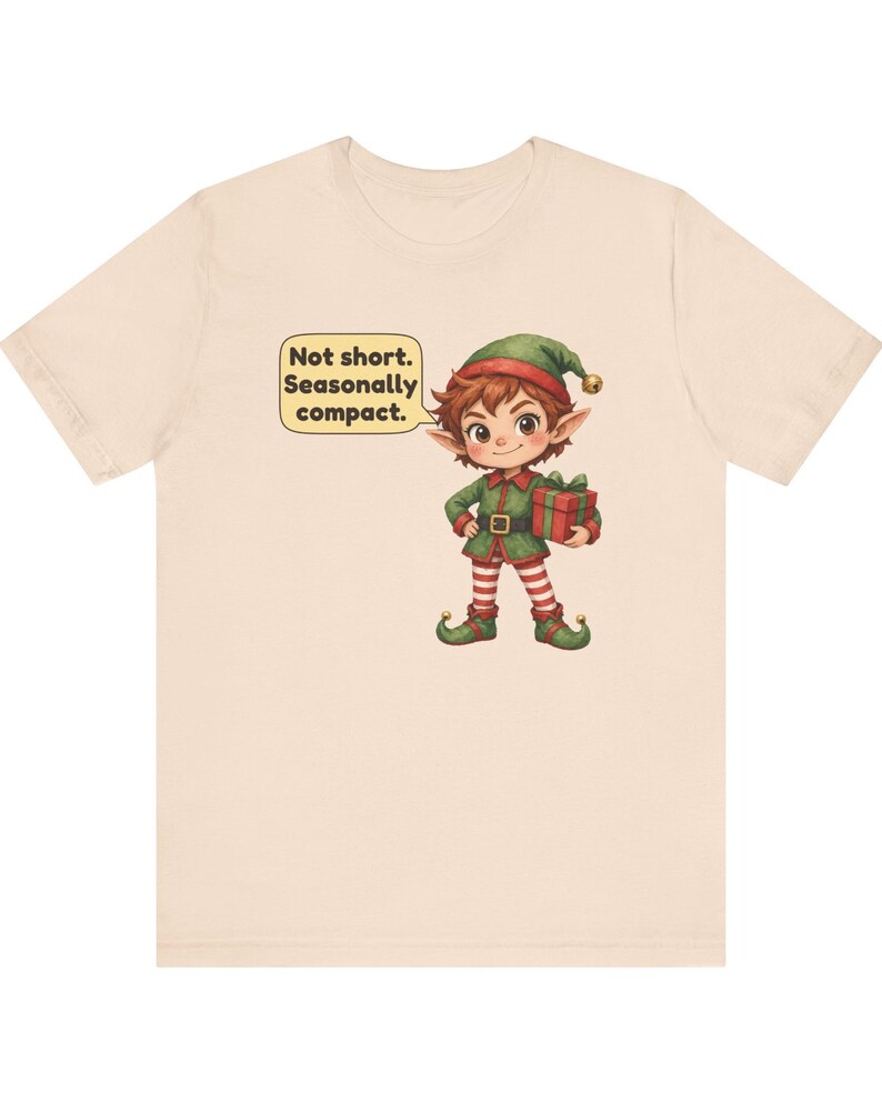 May include: A light beige t-shirt featuring a cartoon elf wearing a green outfit with red and white striped leggings, holding a red gift box. A speech bubble next to the elf contains the text: Not short. Seasonally compact.
