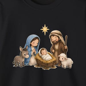 May include: A black long-sleeve shirt featuring a colorful graphic of a nativity scene. The illustration depicts a person in a blue head covering, a person in brown robes with a staff, a baby in a manger, a donkey, a sheep, and a star.