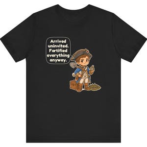 May include: A black t-shirt featuring a cartoon illustration of a historical explorer in a blue coat and tricorn hat, holding a shovel and compass next to a wooden chest. A speech bubble contains the text: Arrived uninvited. Fortified everything anyway.