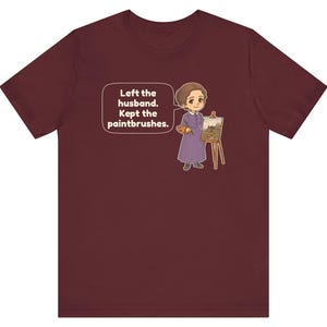 May include: A maroon short-sleeve t-shirt featuring a cartoon illustration of a person in a purple dress standing by an easel with a paintbrush and palette. A speech bubble contains the text: Left the husband. Kept the paintbrushes.
