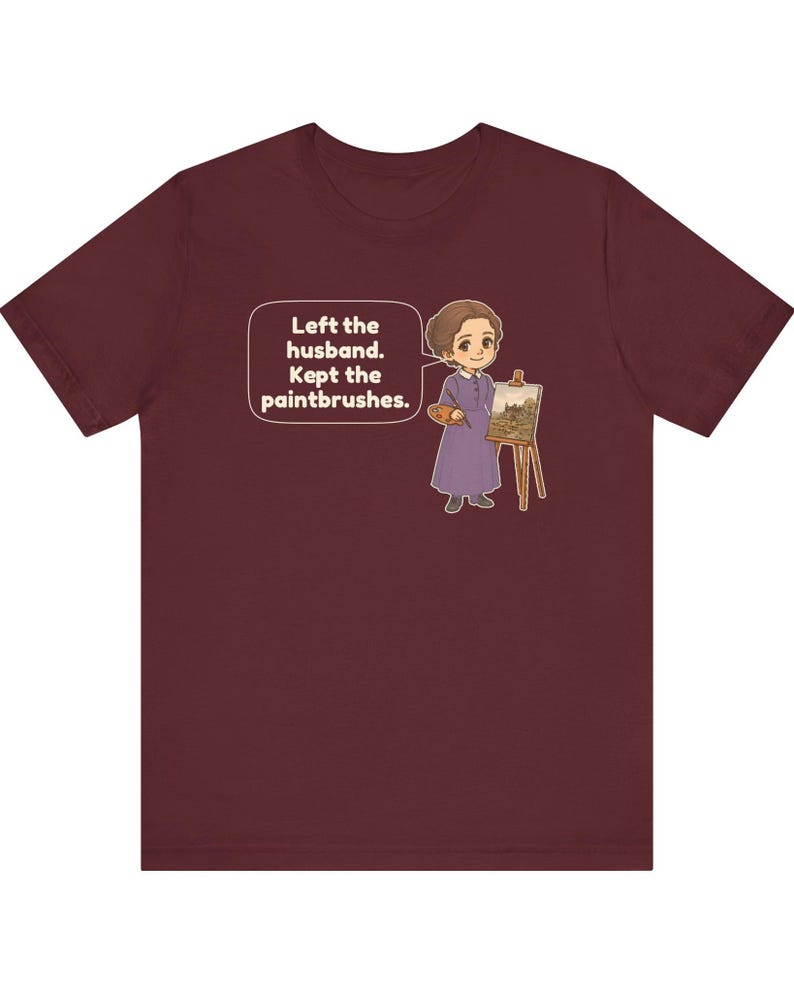 May include: A maroon short-sleeve t-shirt featuring a cartoon illustration of a person in a purple dress standing by an easel with a paintbrush and palette. A speech bubble contains the text: Left the husband. Kept the paintbrushes.