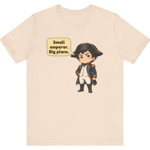 May include: A light beige t-shirt featuring a cartoon illustration of a historical figure in a blue military coat, white breeches, and a black bicorne hat. A yellow speech bubble next to the character contains the text: Small emperor. Big plans.