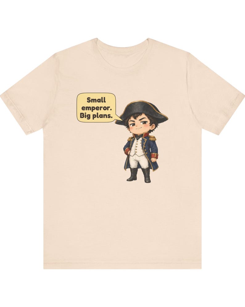 May include: A light beige t-shirt featuring a cartoon illustration of a historical figure in a blue military coat, white breeches, and a black bicorne hat. A yellow speech bubble next to the character contains the text: Small emperor. Big plans.