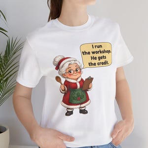 K&ouml;nnte beinhalten: Ein wei&szlig;es kurz&auml;rmeliges T-Shirt mit einer Grafik einer &auml;lteren Frau in einem rot-wei&szlig;en Festtagsoutfit, die einen Holzl&ouml;ffel und ein Klemmbrett h&auml;lt. Eine gelbe Sprechblase enth&auml;lt den Text: I run the workshop. He gets the credit.