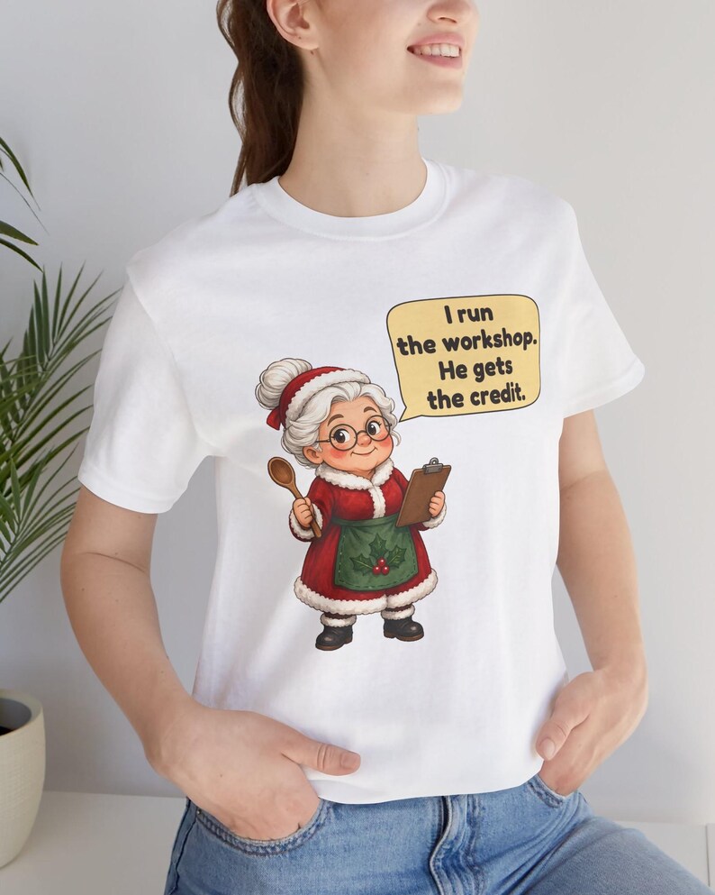 K&ouml;nnte beinhalten: Ein wei&szlig;es kurz&auml;rmeliges T-Shirt mit einer Grafik einer &auml;lteren Frau in einem rot-wei&szlig;en Festtagsoutfit, die einen Holzl&ouml;ffel und ein Klemmbrett h&auml;lt. Eine gelbe Sprechblase enth&auml;lt den Text: I run the workshop. He gets the credit.