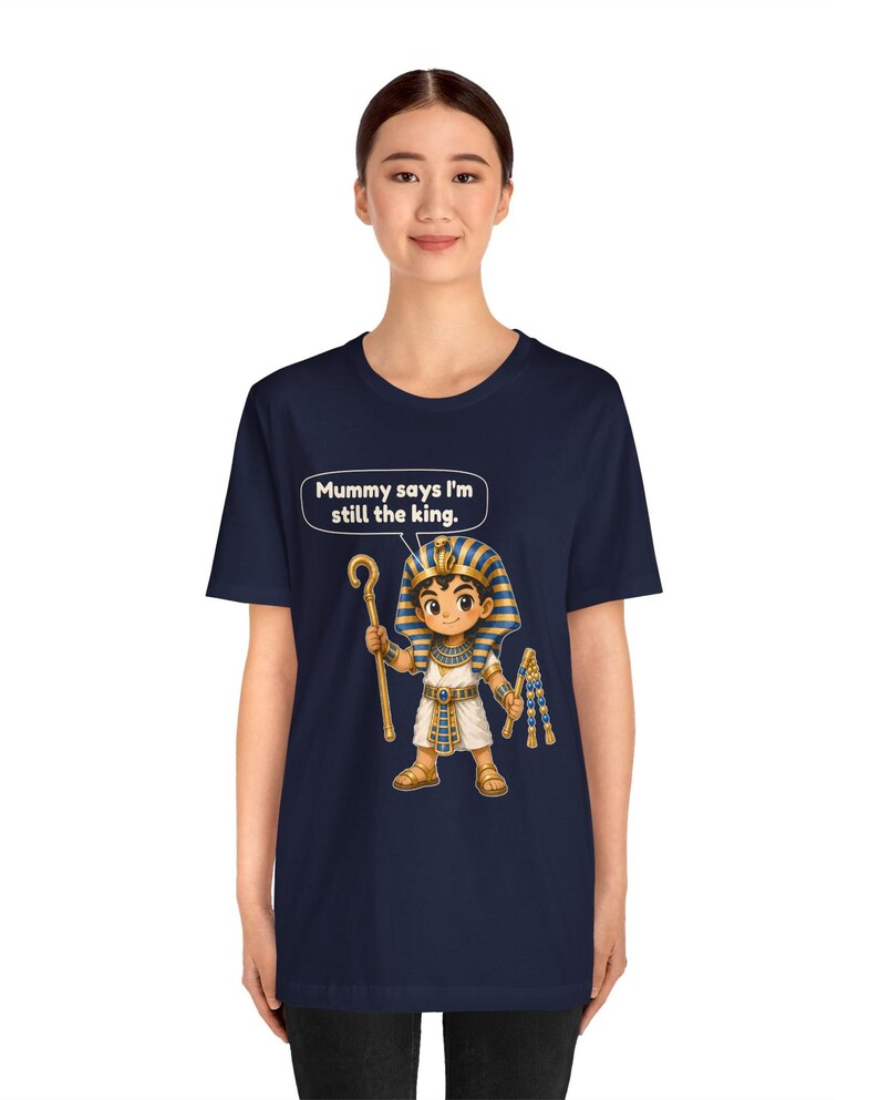 Może przedstawiać: Granatowy t-shirt z grafiką przedstawiającą faraona z kresk&oacute;wki trzymającego laskę i bicz. Nad postacią znajduje się dymek z tekstem: Mummy says I'm still the king. Faraon ma na głowie niebiesko-złote nakrycie głowy w paski.