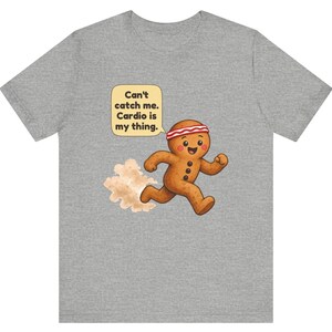 May include: A heather grey t-shirt featuring a cartoon gingerbread person running with a red and white headband. A speech bubble above the character contains the text: Can't catch me. Cardio is my thing. A small cloud of dust follows the figure.